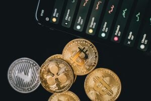fotografía de los tokens de las criptomonedas más famosas, como bitc,eth,ripple, al lado de un celular con los precios de las criptomonedas