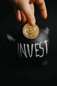 Fotografia de una mano sosteniendo un token de btc y atrás una alcancia de cochino con la palabra "invest" en ella, analizando por que no invertir en criptomonedas